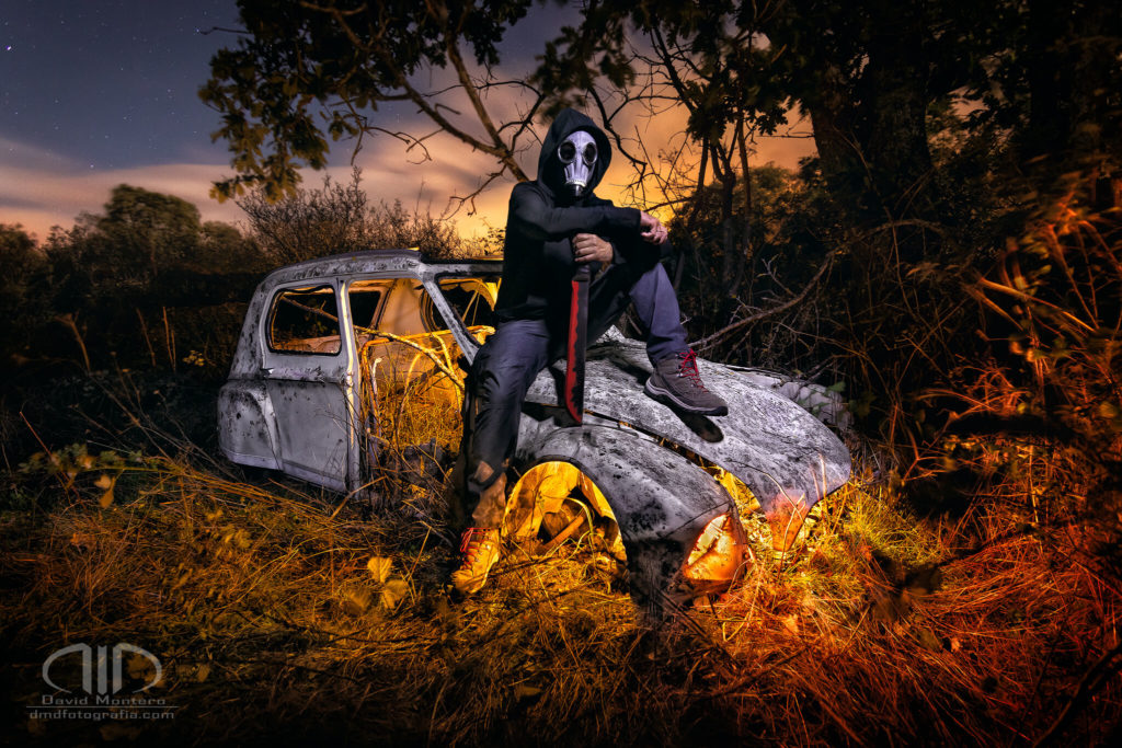 Personaje apocalíptico con máscara de gas y capucha ostiene un machete sobre un coche abandonado