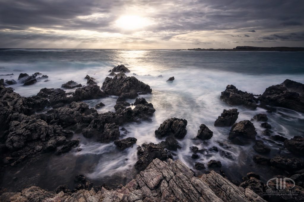 Amanecer en la costa de Menorca. Olas rompiendo contra las piedras mientras sale el sol entre las nubes